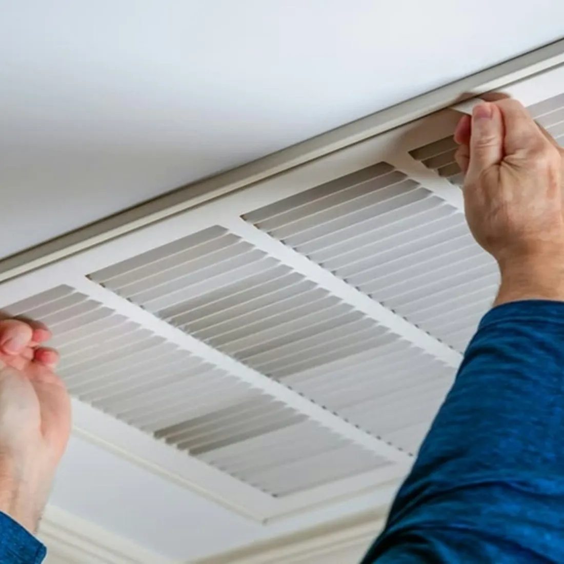 Two hands lifting a white louvred return-air vent grille away from a ceiling