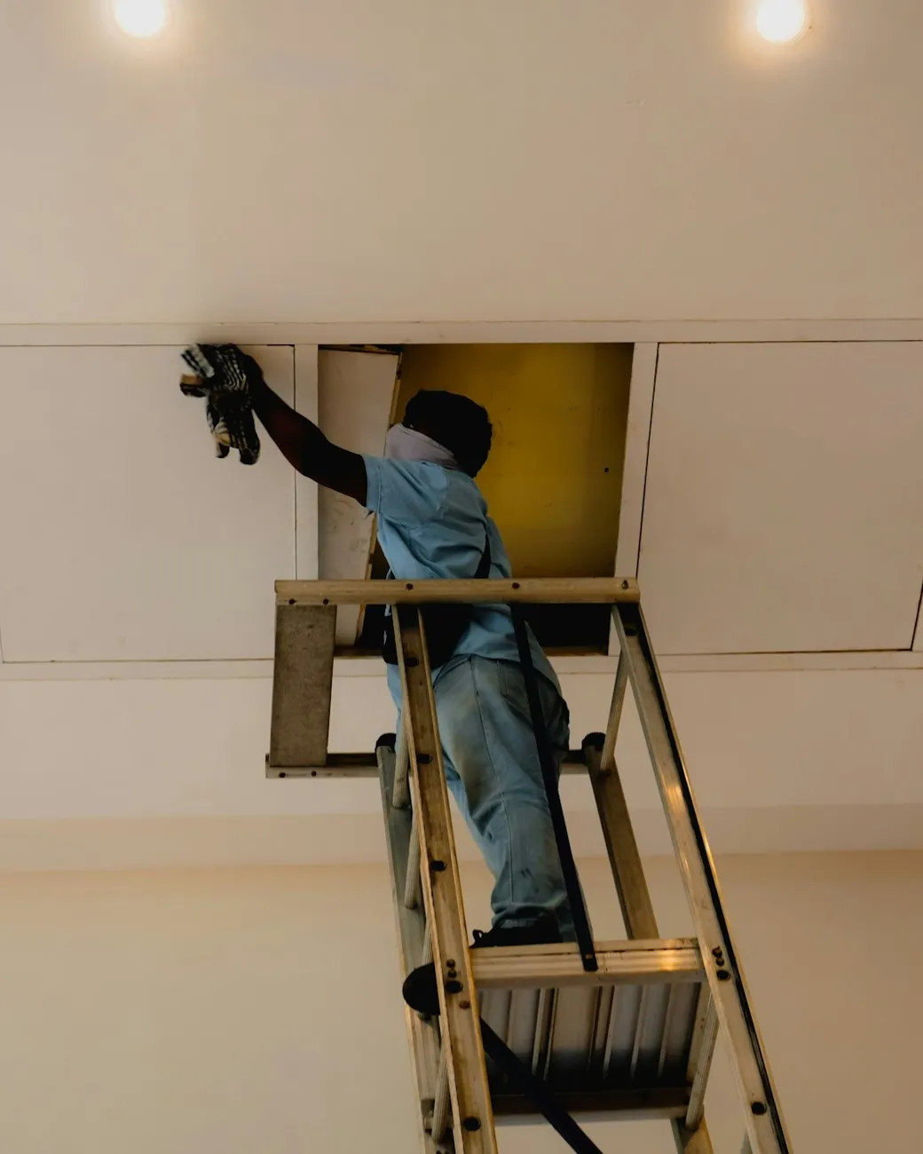 Technician in a light-blue short-sleeve work shirt on a platform ladder, reaching into an open ceiling access hatch