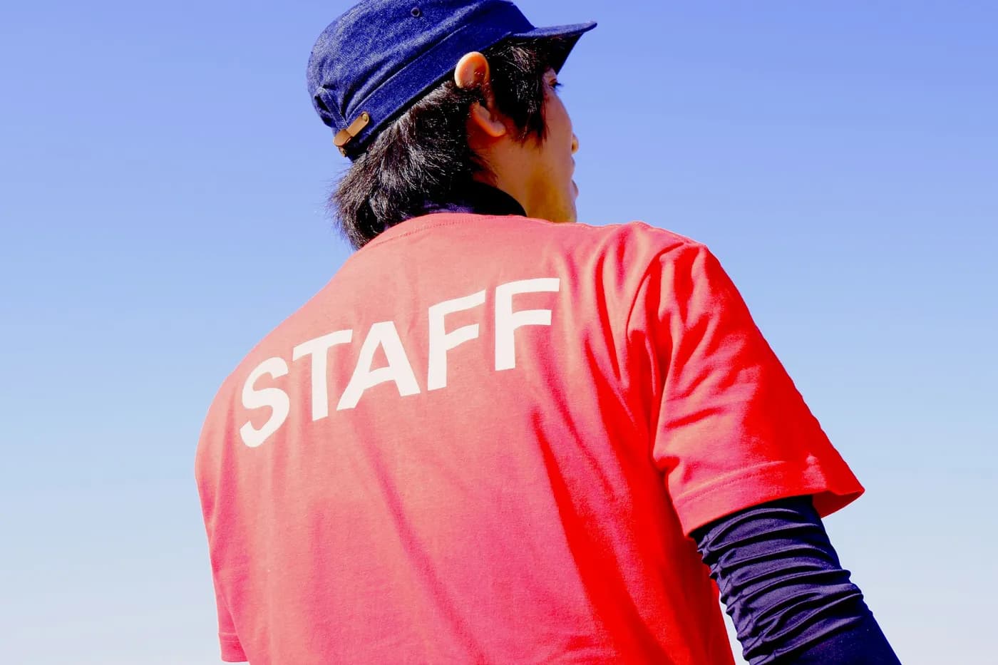 Person seen from behind in a red t-shirt with STAFF printed in large white letters across the back, against a blue sky