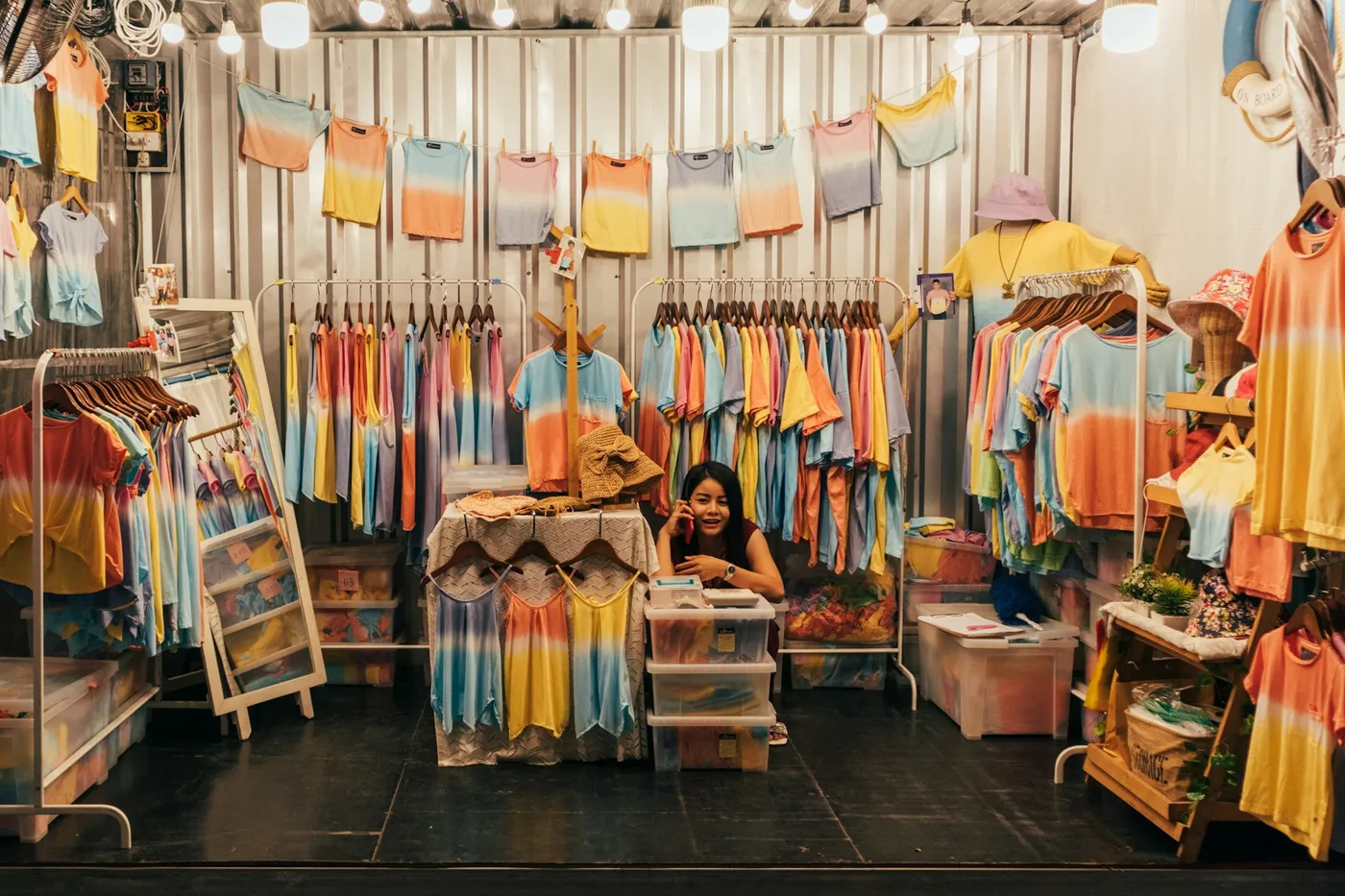Pop-up clothing stall lit by bare bulbs, with rows of pastel dip-dyed t-shirts on rails and a vendor behind the counter