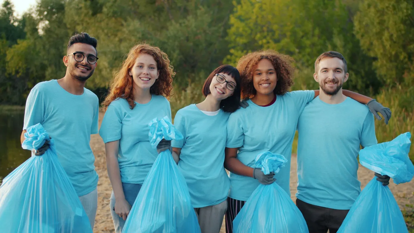 Five volunteers in matching sky-blue t-shirts and work gloves holding full garbage bags on a lakeside path