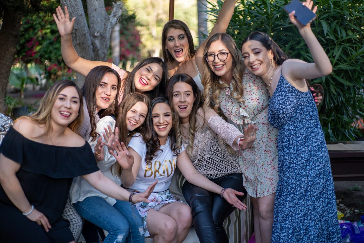 About nine women laughing and waving in a leafy garden, the bride-to-be in the centre wearing a white t-shirt with gold BRIDE lettering
