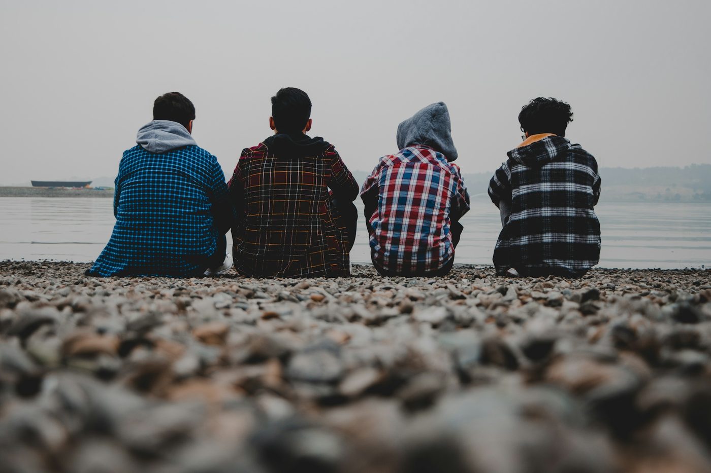 Four friends seen from behind sitting on a pebble beach facing a calm grey lake, in plaid flannel shirts, one with a grey hood up
