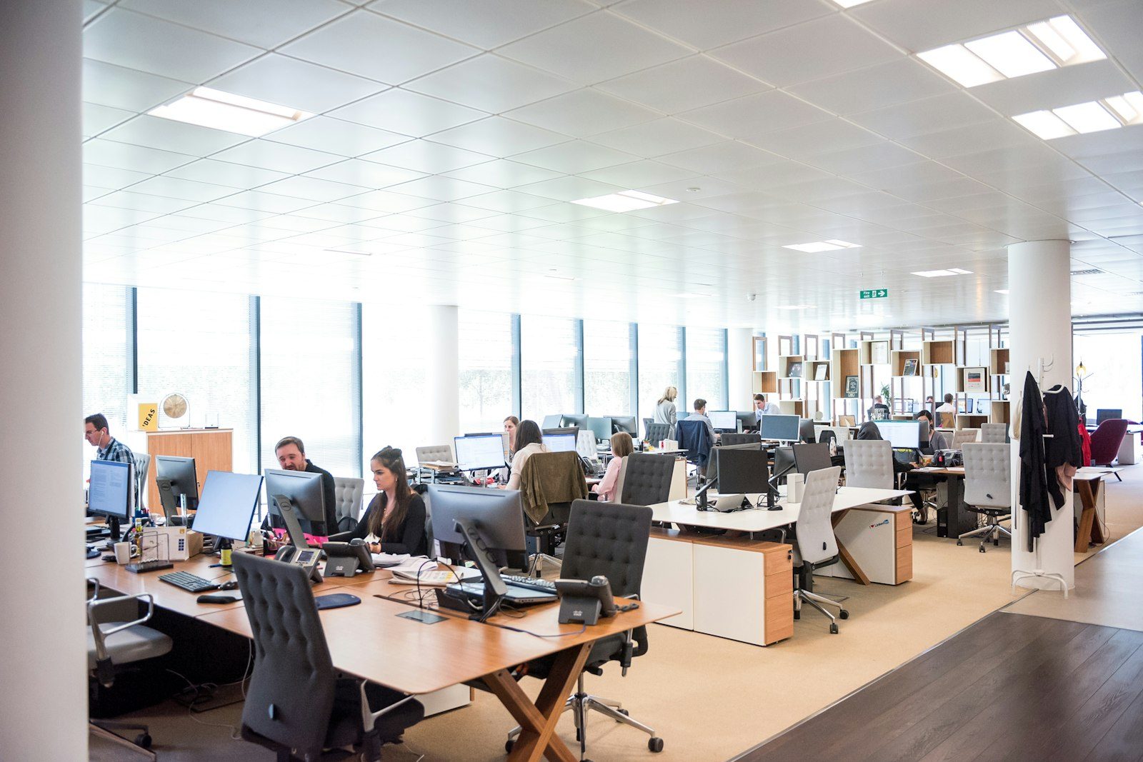 A bright open-plan office with long rows of desks and monitors and people at work