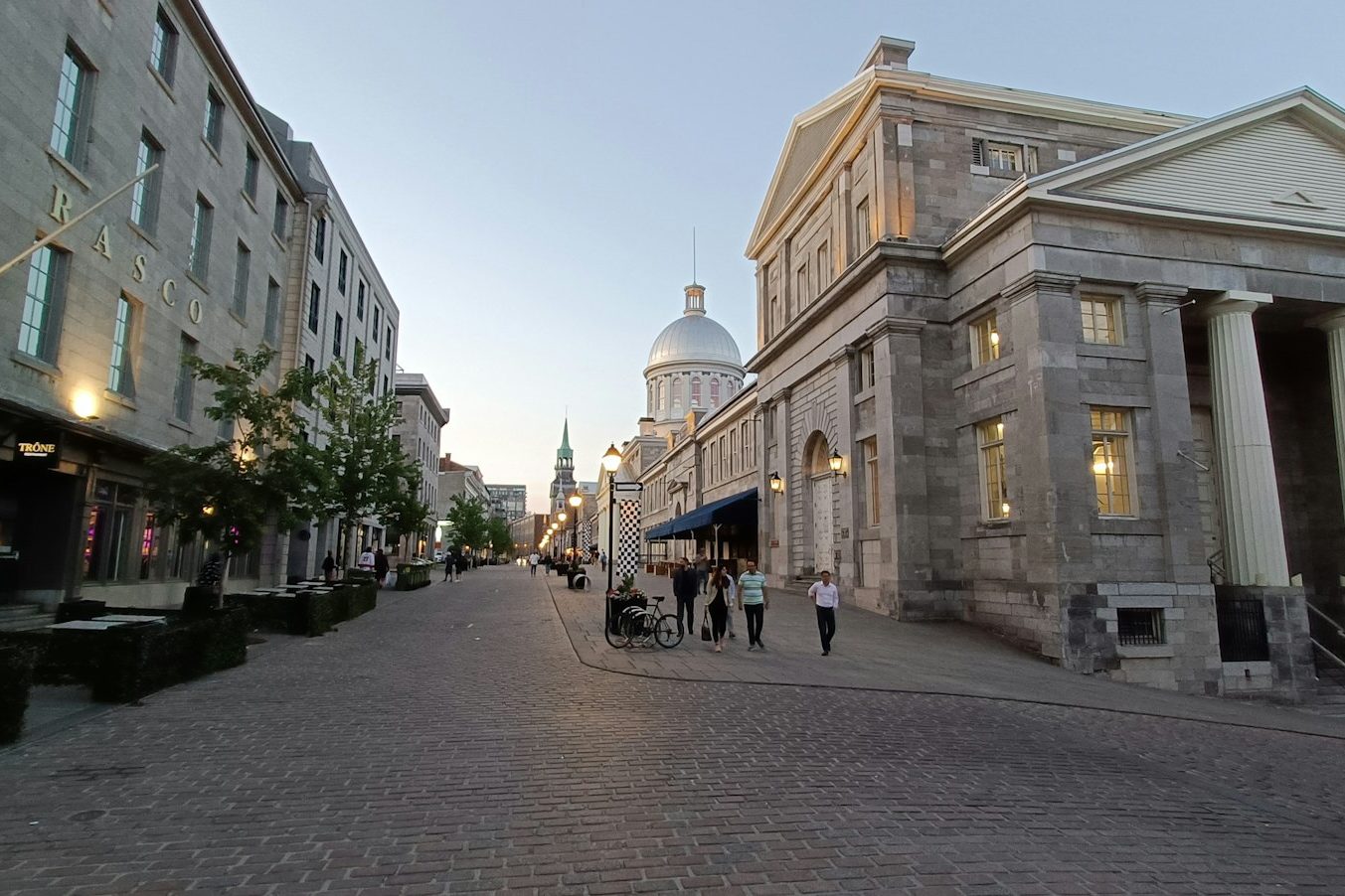 A cobblestone street in Old Montreal at dusk, lined with grey stone buildings, the silver dome of Bonsecours Market at the end