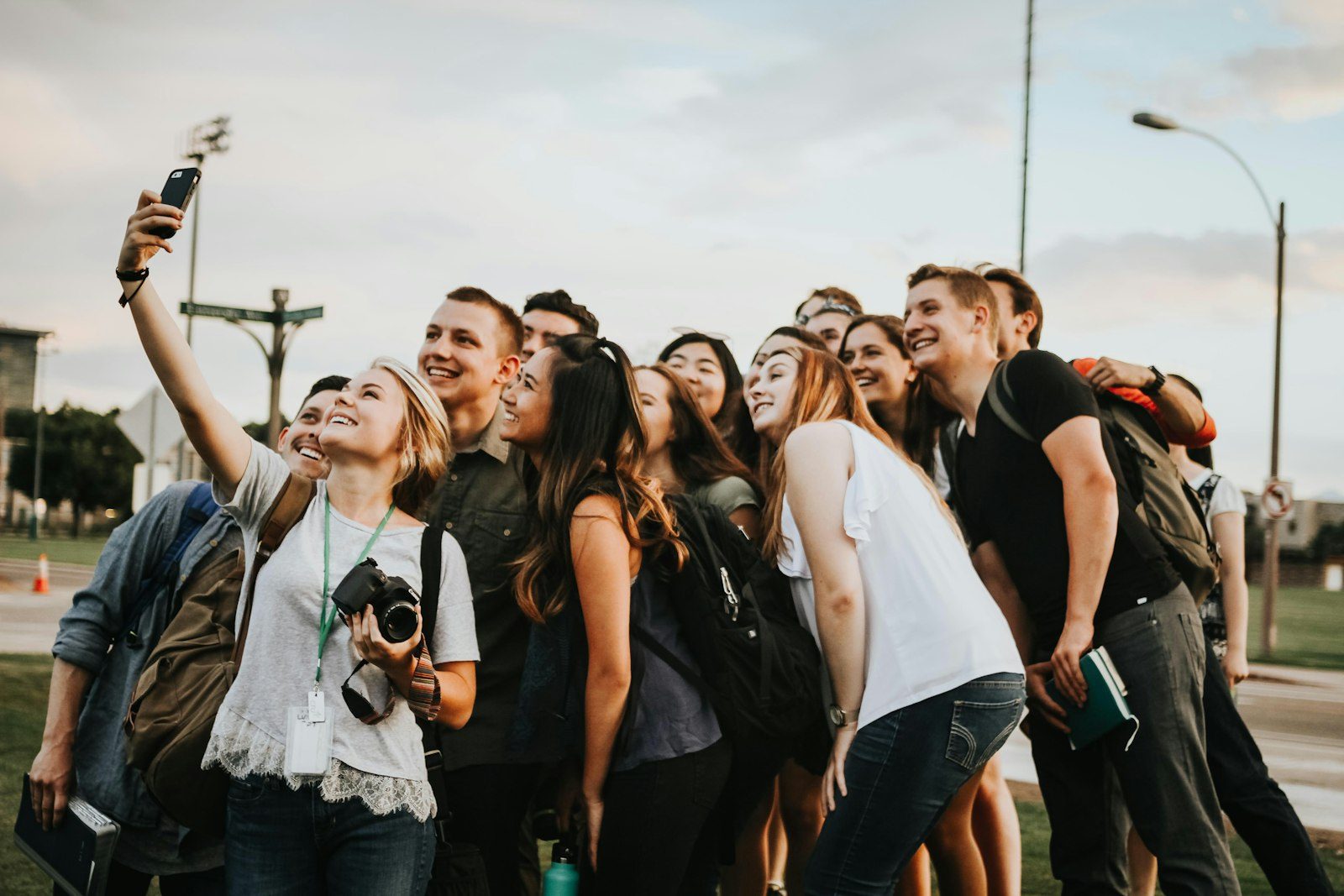 A group of young people crowding together for a selfie outdoors, one wearing a lanyard and a camera