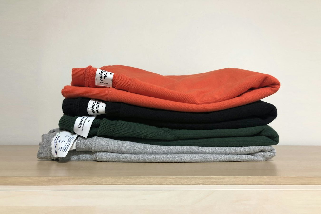 Four folded fleece sweatshirts in orange, black, dark green and grey stacked on a pale wooden shelf