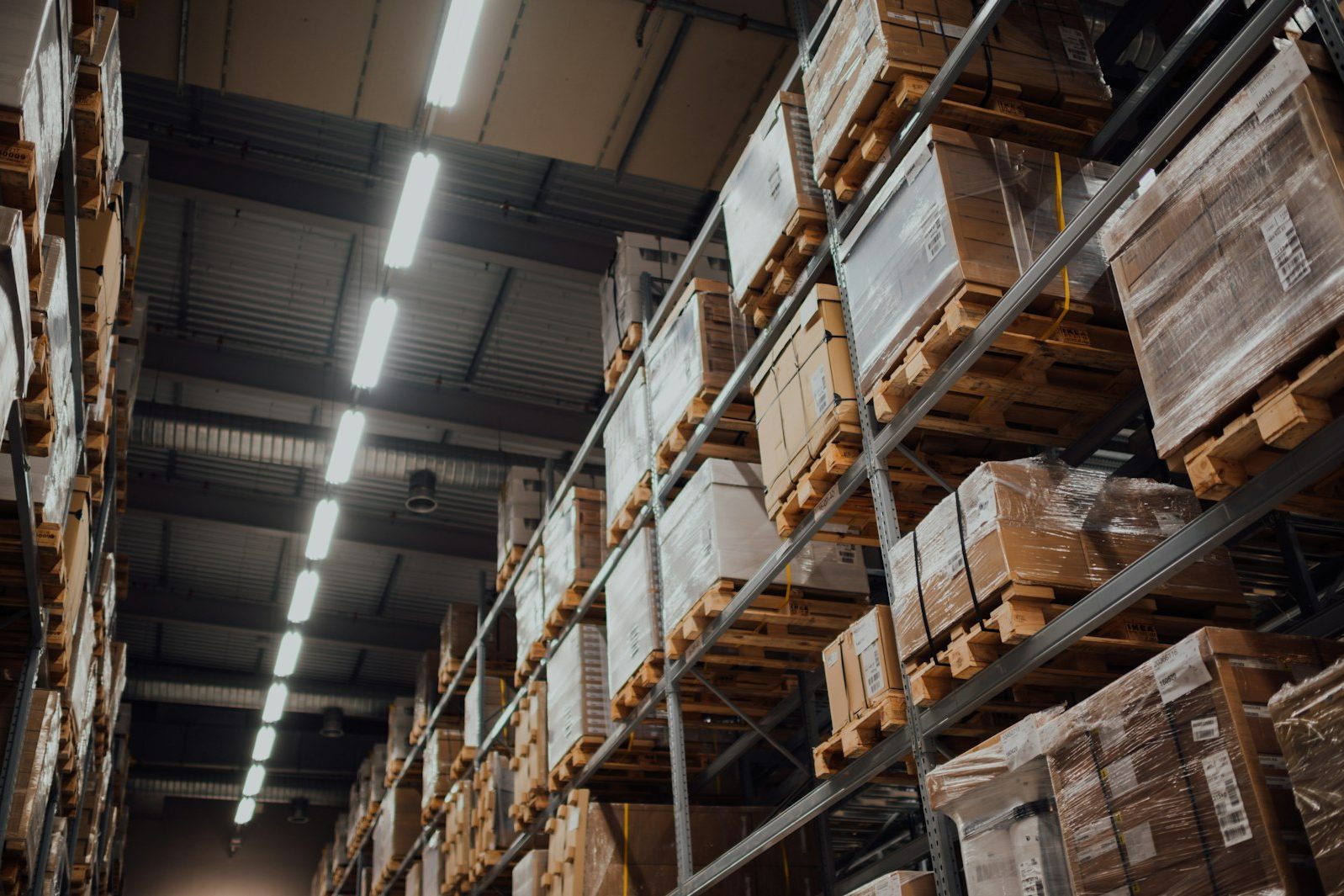 Pallets of cardboard cartons stacked high on warehouse racking under long ceiling lights