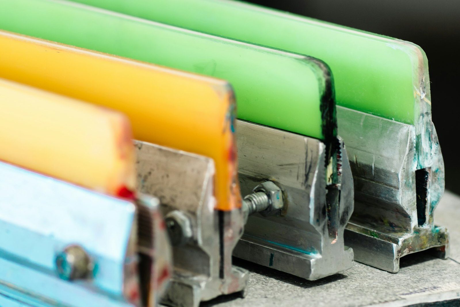 Close-up of screen-printing squeegees with yellow, green and blue rubber blades clamped in metal handles