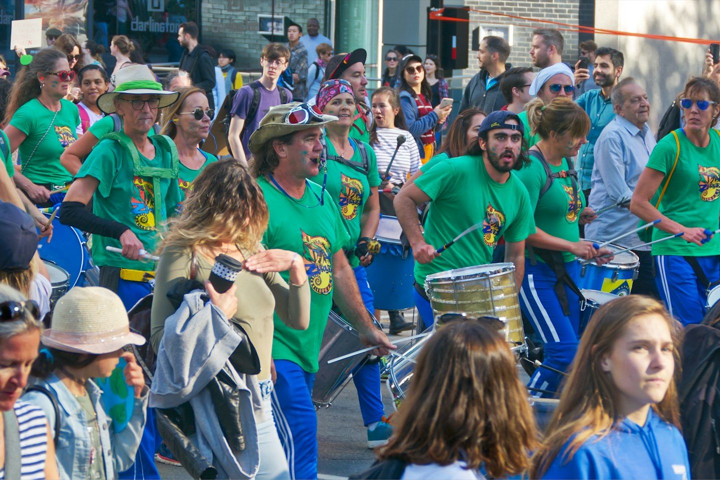 Drummers in matching green t-shirts marching through a crowded city street during a parade