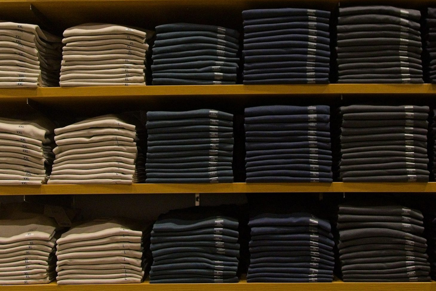Shelves of folded t-shirts stacked by colour in cream, navy and charcoal, with size stickers on the edges of the piles