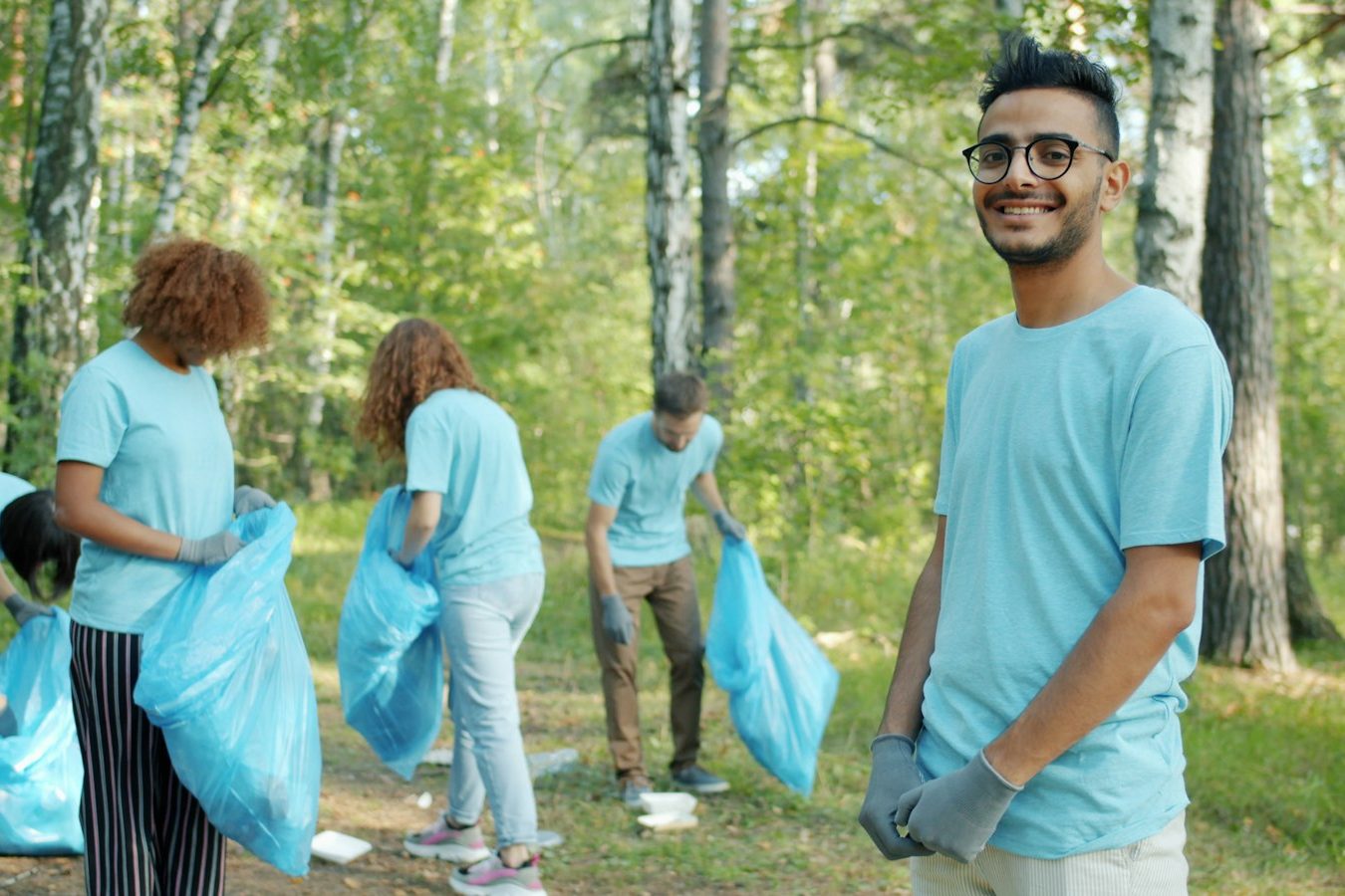 Volunteers in matching light-blue t-shirts picking up litter in a wooded park, one smiling at the camera