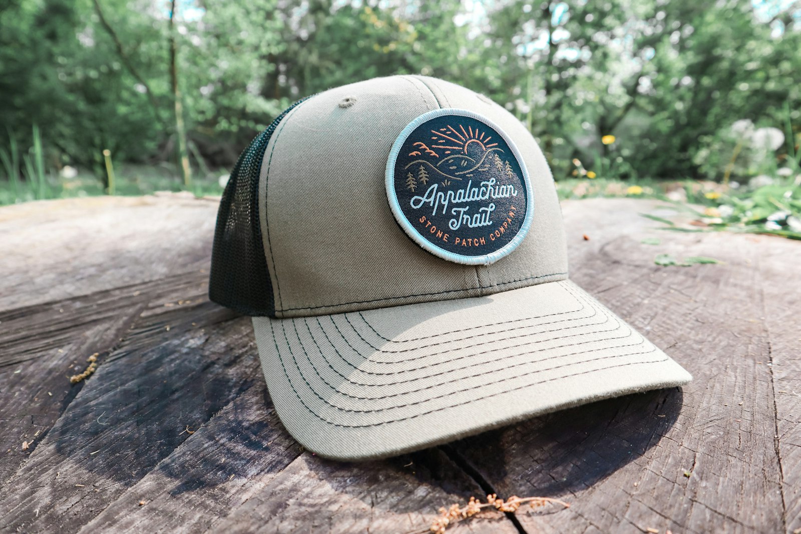 An olive and black trucker cap with a round embroidered patch sitting on a wooden surface