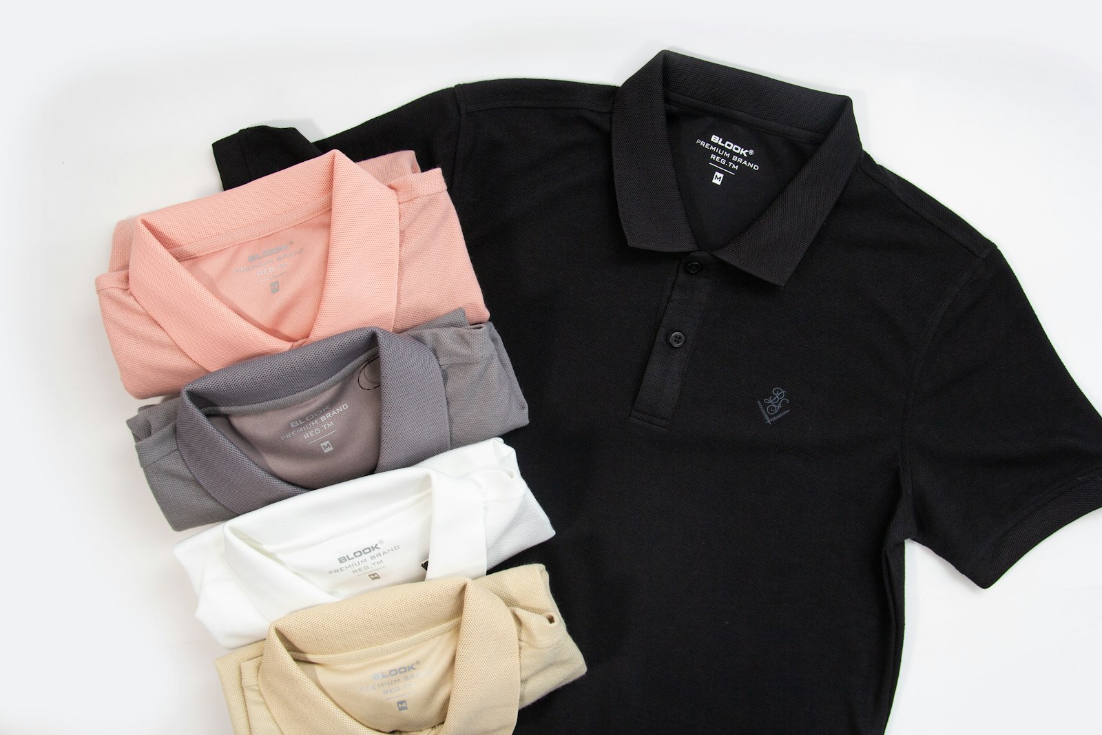 Folded polo shirts in black, coral, grey, white and tan laid out flat on a white surface