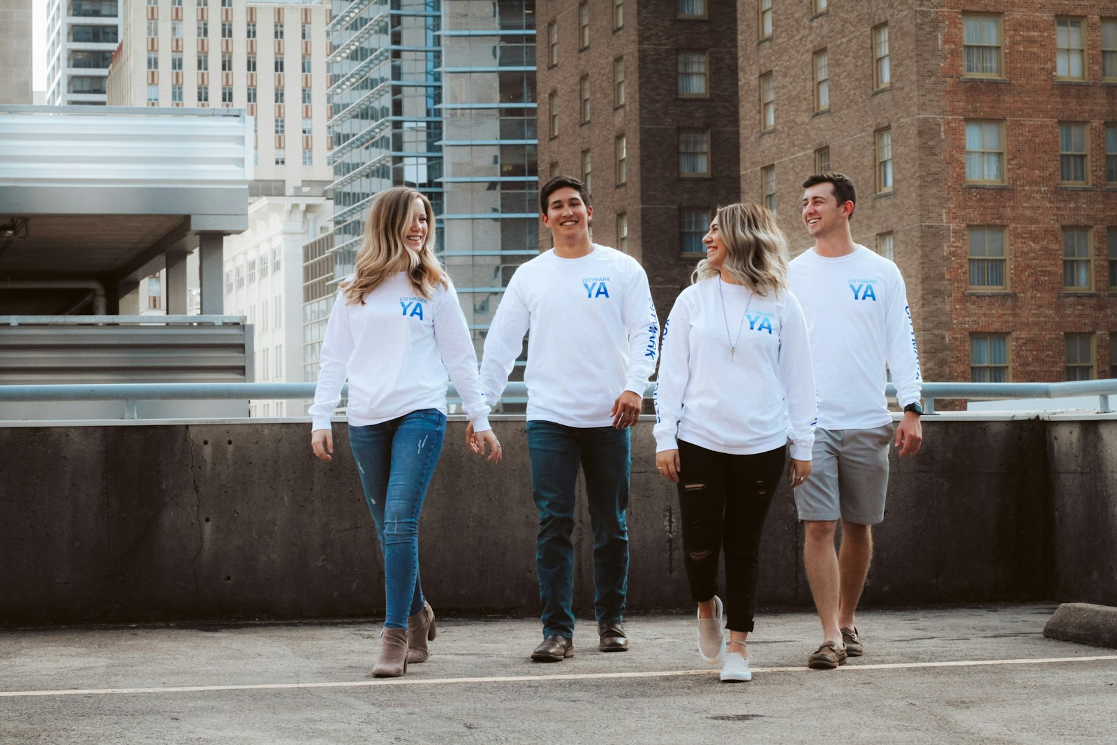Four coworkers in matching white long-sleeve shirts with a small chest logo walking together on a downtown street