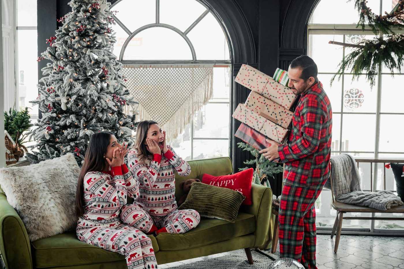 Two women in matching red-and-white holiday pyjamas on a green sofa as a man in red tartan pyjamas carries in a stack of gifts beside a Christmas tree