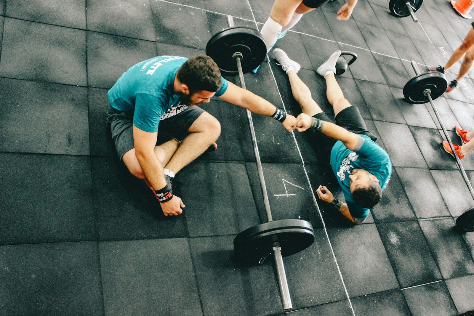 Two people in matching teal t-shirts stretching on a black gym floor beside a loaded barbell, seen from above