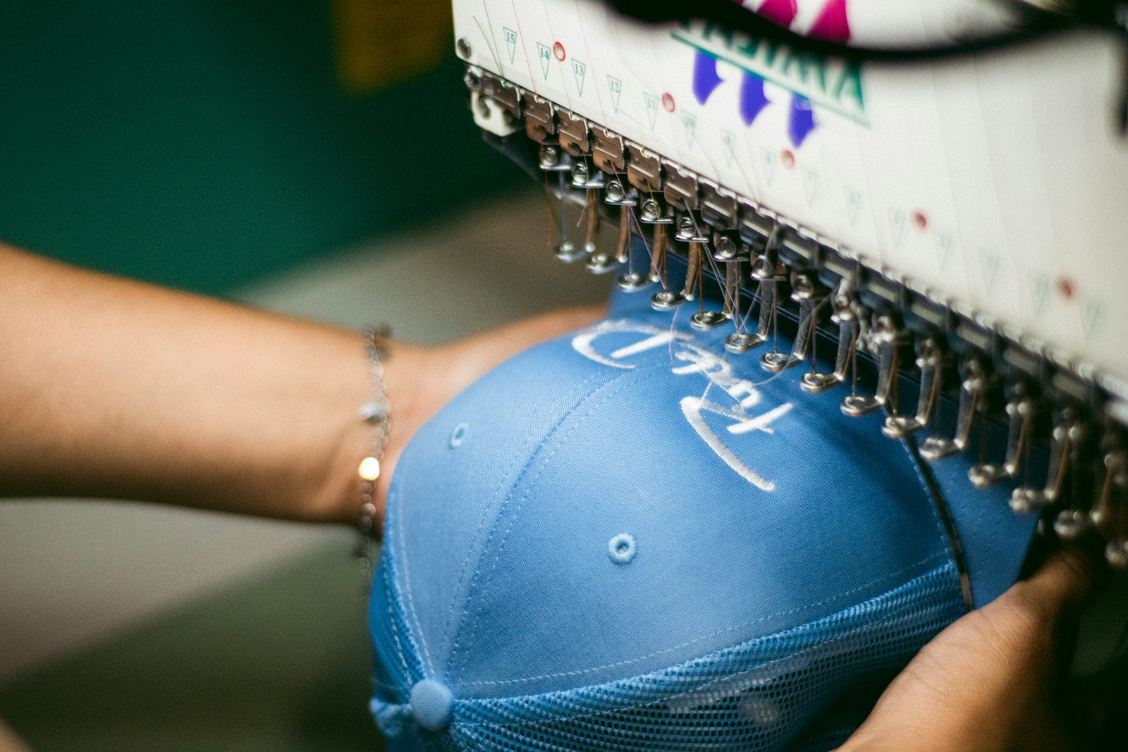 A blue cap held in the hoop of an embroidery machine as a white script logo is stitched onto the front