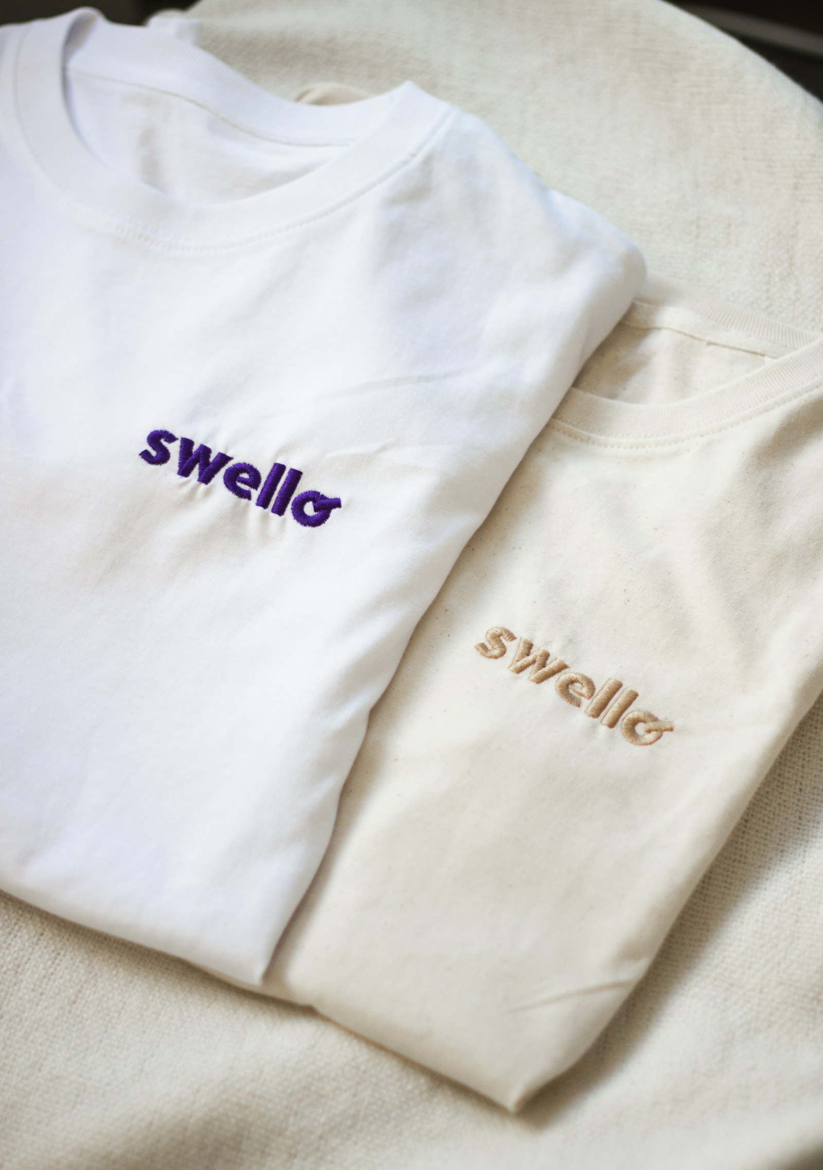 Two folded garments in white and cream, each with a small embroidered wordmark on the chest