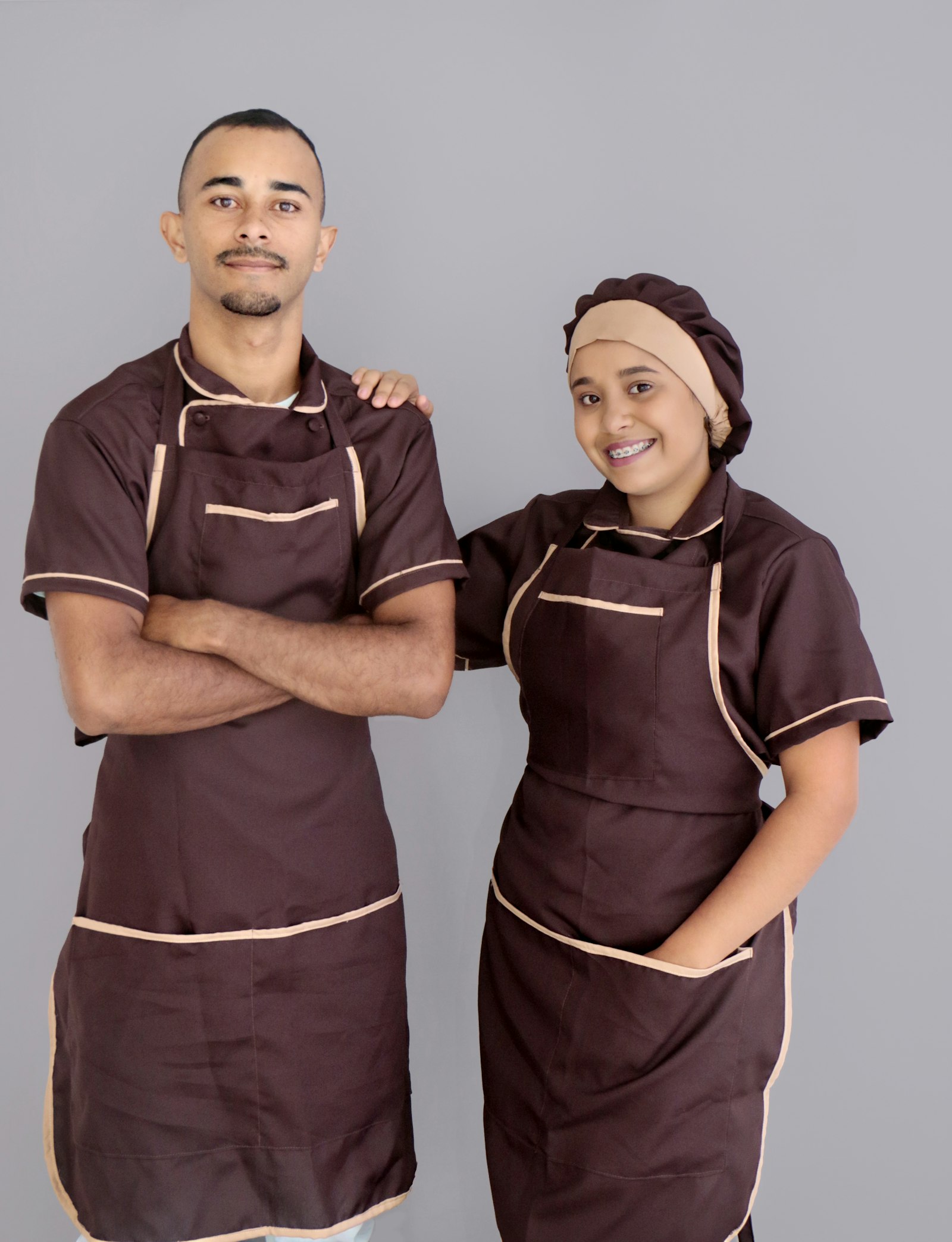 Two restaurant workers in matching brown aprons with cream piping, arms crossed, against a grey backdrop