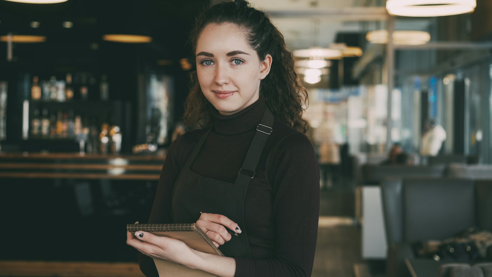 A server in a dark apron holding a tablet inside a restaurant with a bar in the background