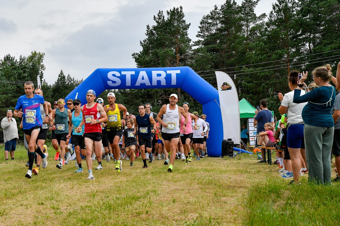 A pack of runners in singlets and t-shirts with race bibs leaving a blue inflatable START arch onto a grassy field lined with pines