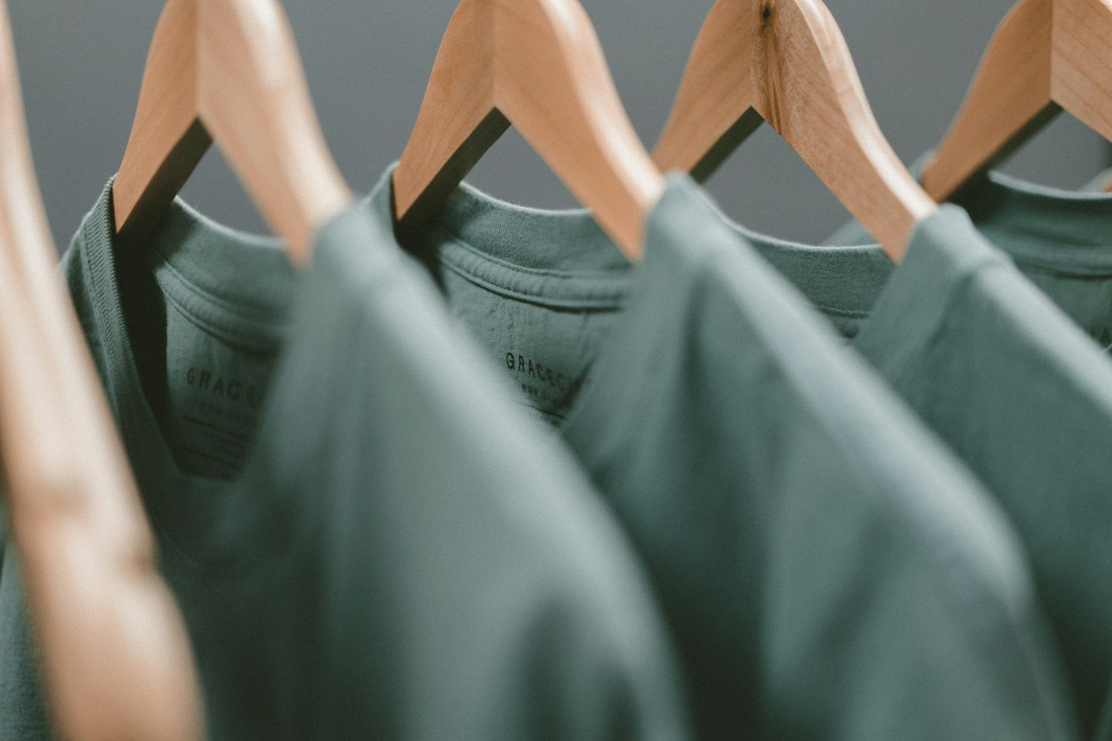 A row of sage-green t-shirts on wooden hangers, each with the same small print on the chest