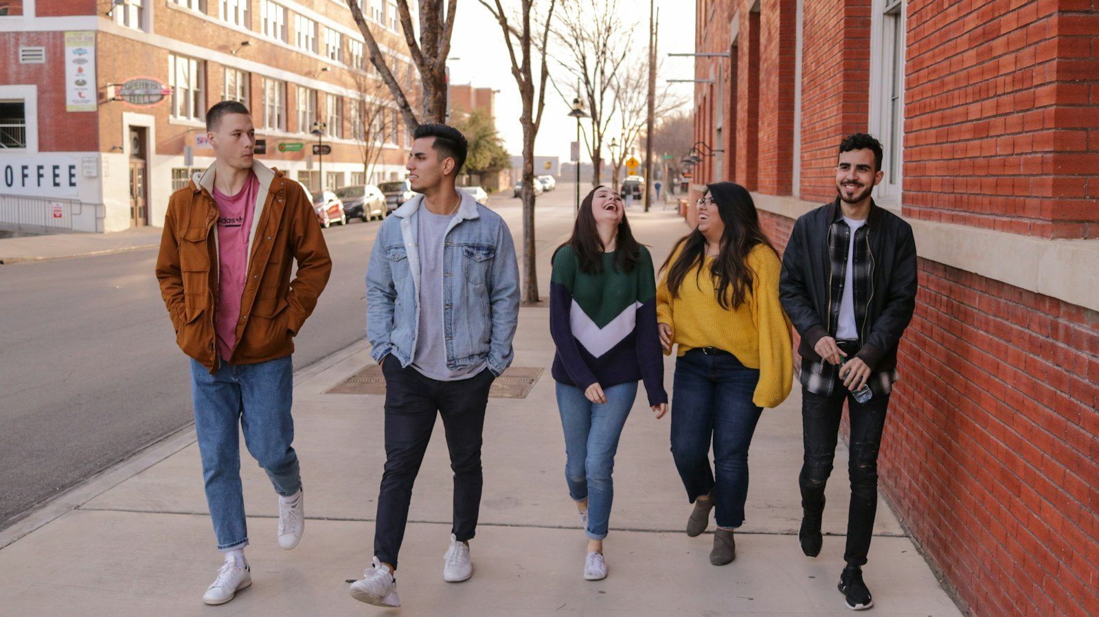 Five teenagers in jackets and hoodies walking side by side along a sidewalk past a brick school building