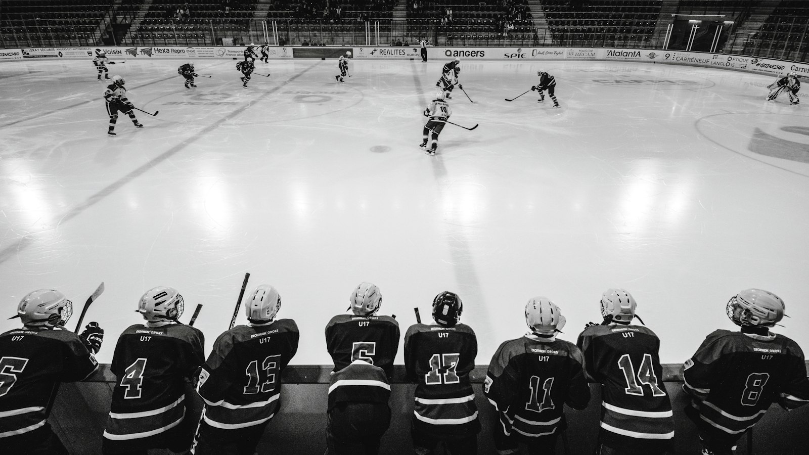 Black-and-white photo of hockey players seated on the bench in numbered jerseys, watching play on the ice