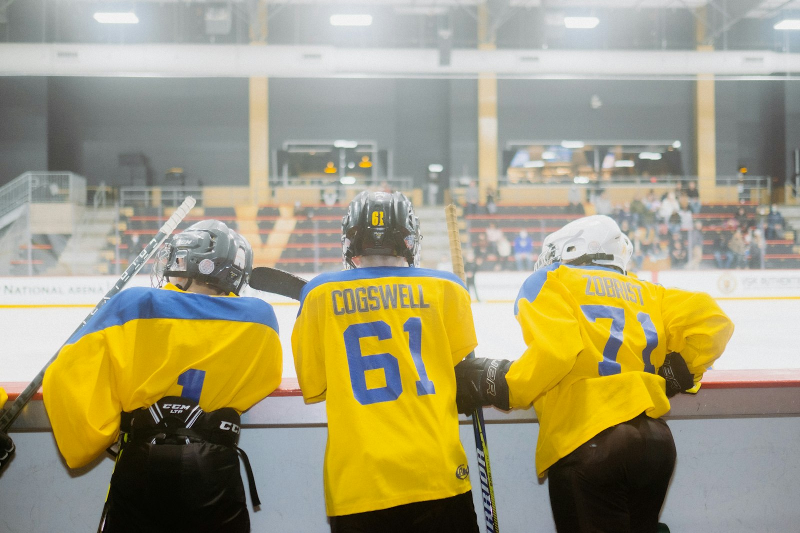 Three young hockey players in yellow and blue jerseys with names and numbers on the back, leaning on the boards at an arena