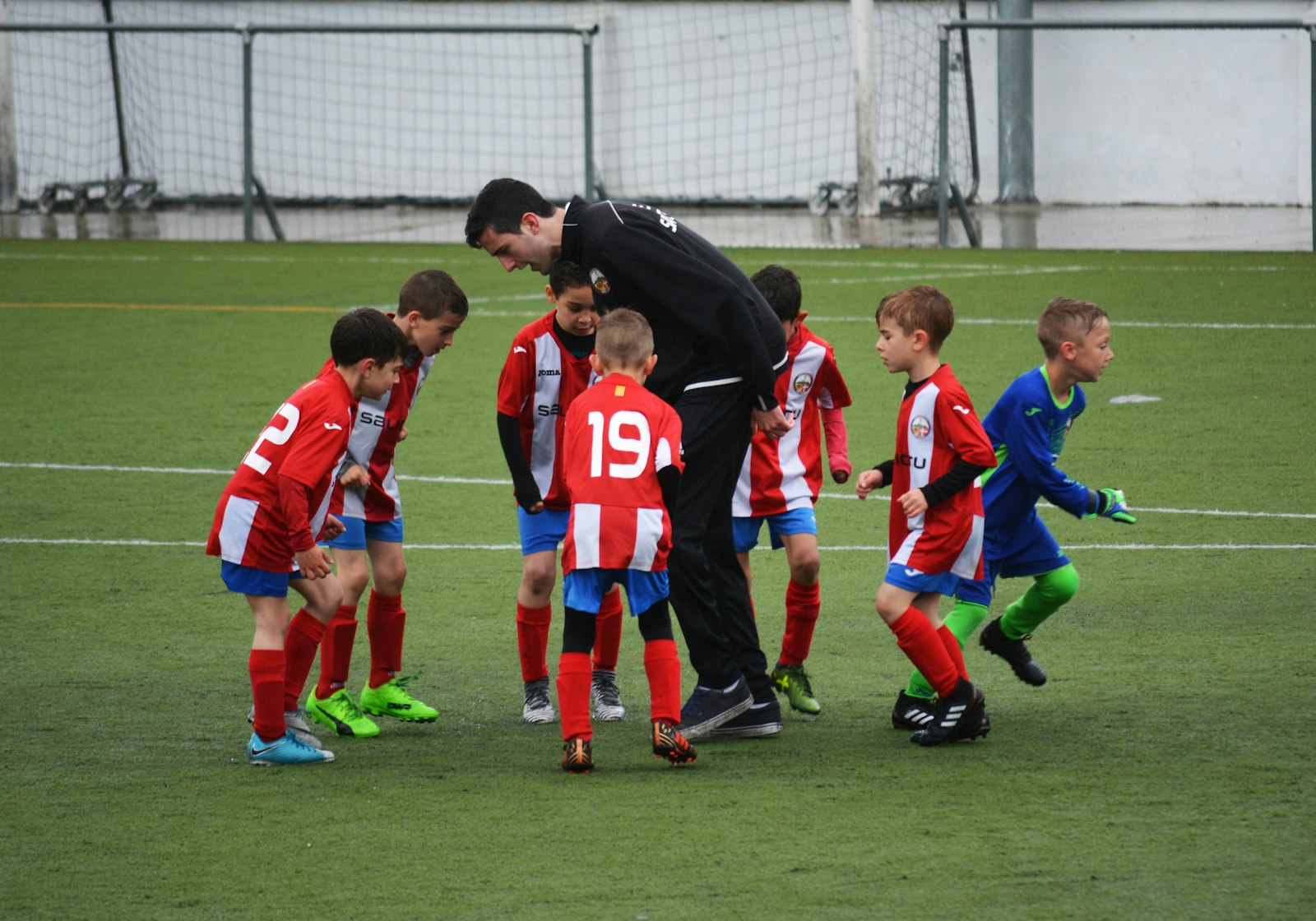 A coach crouched among young players in red-and-blue and green kits during a huddle on artificial turf