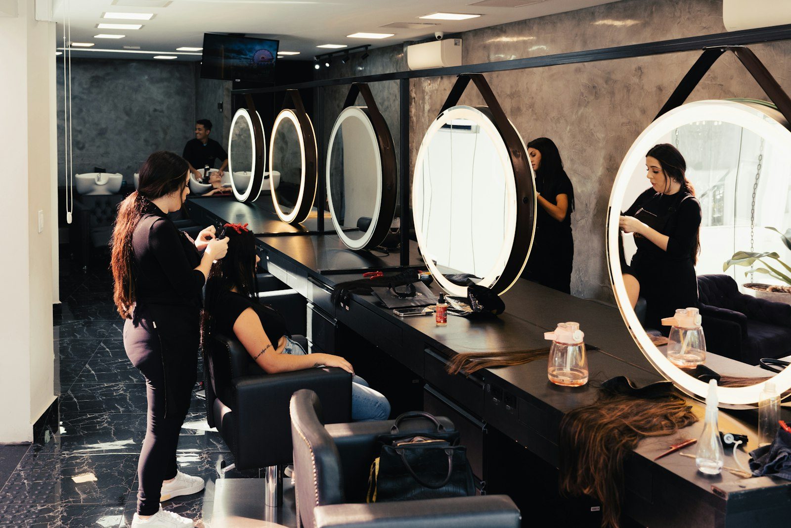 A hair salon with stylists in matching black working at a row of round mirrors along a dark wall