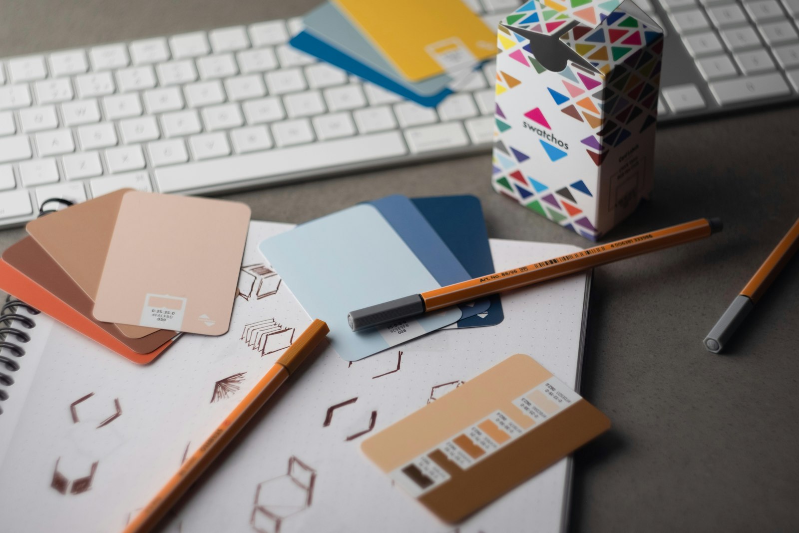 A design desk with a keyboard, sketch pad, pencils and fanned-out colour swatch guides