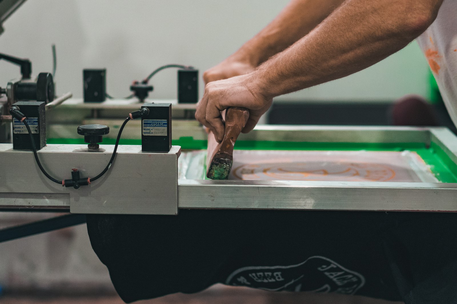 A printer pulling a squeegee across a screen on a flatbed press with a black t-shirt loaded underneath