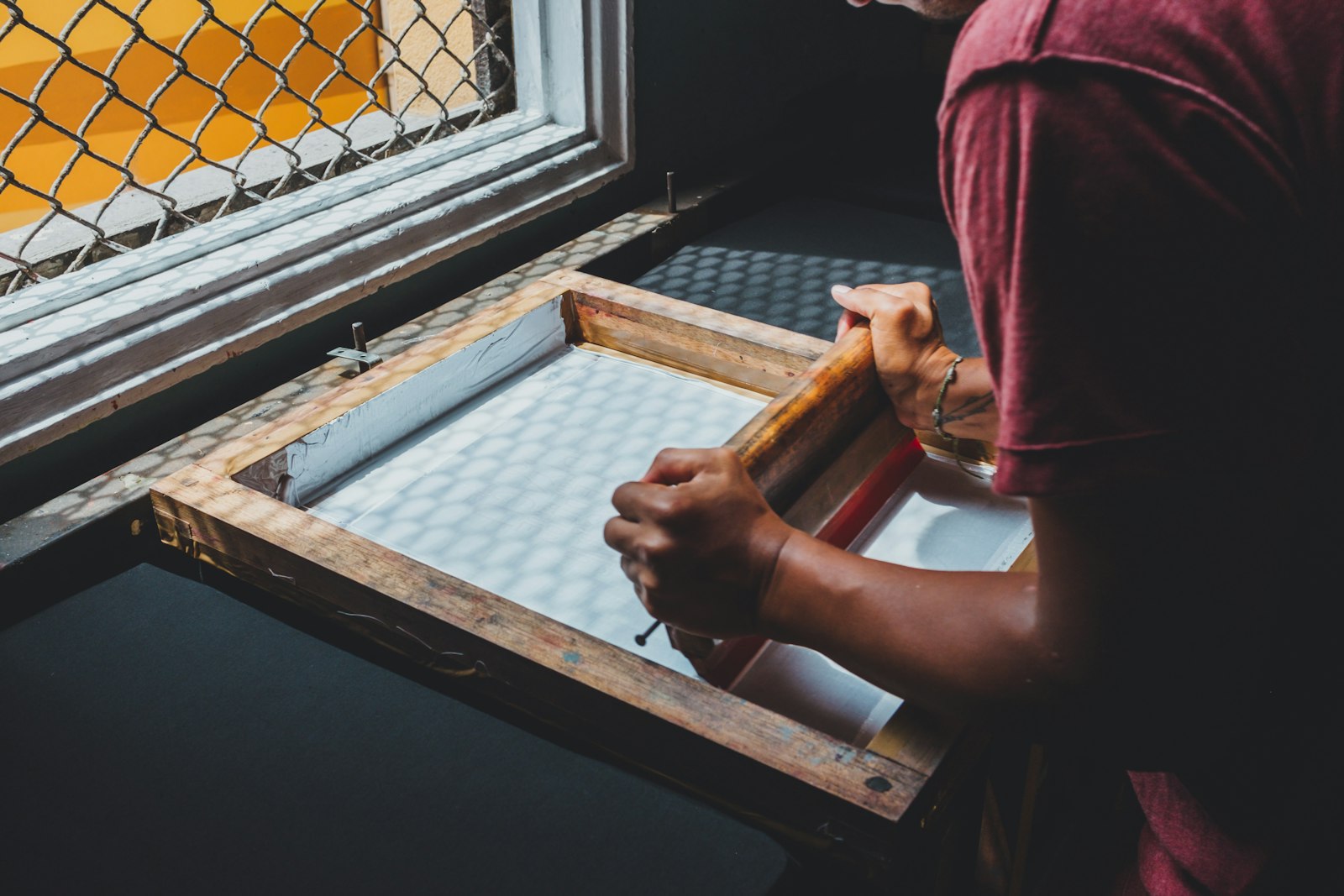 A printer pulling a squeegee of ink across a wooden screen frame on a print bench beside a window