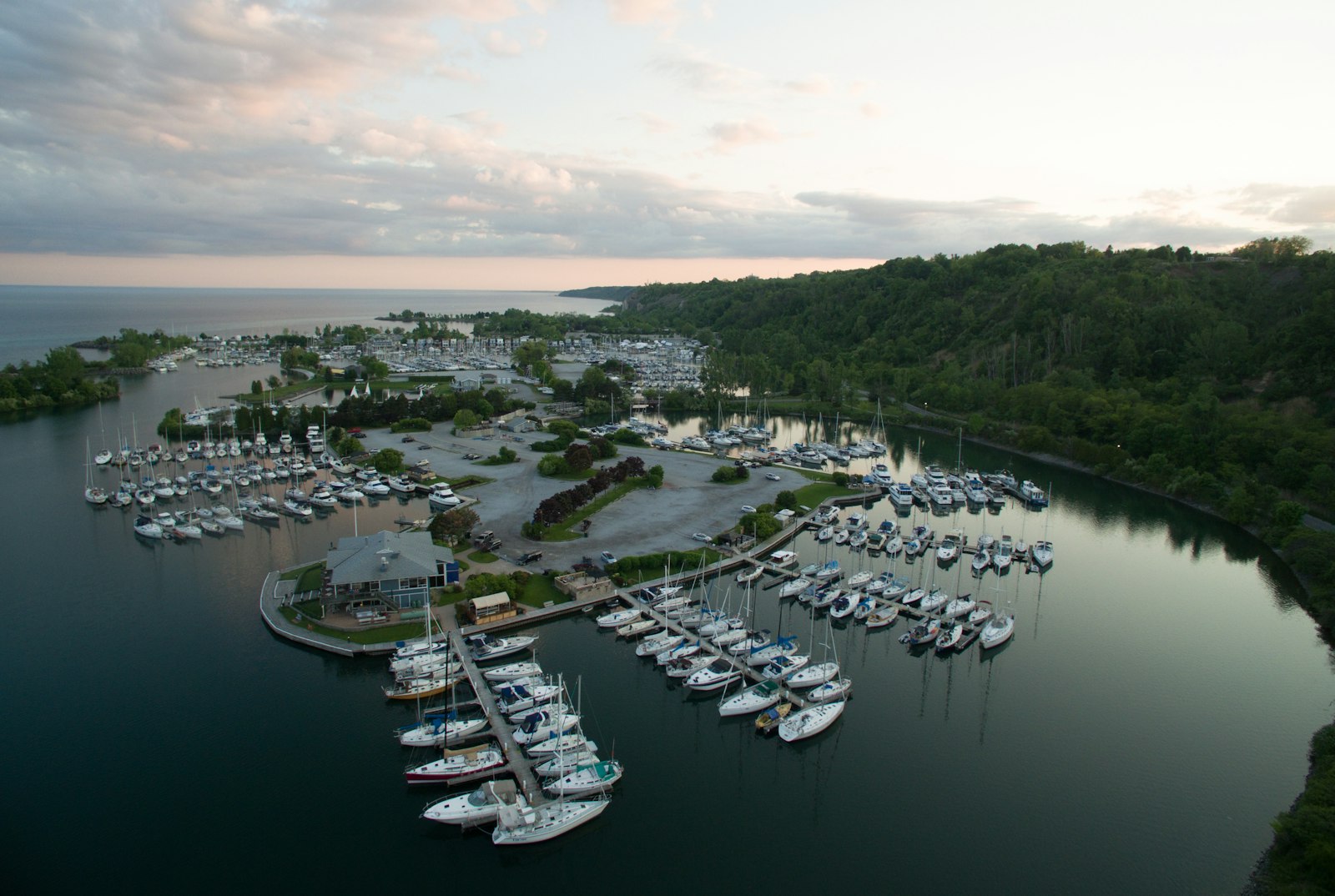 Aerial view at dusk of a small marina full of sailboats tucked behind a wooded shoreline on a lake
