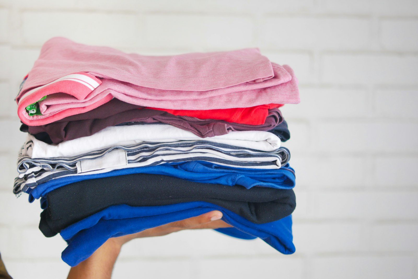 A stack of folded t-shirts in pink, red, white, grey, blue and black held up against a white brick wall