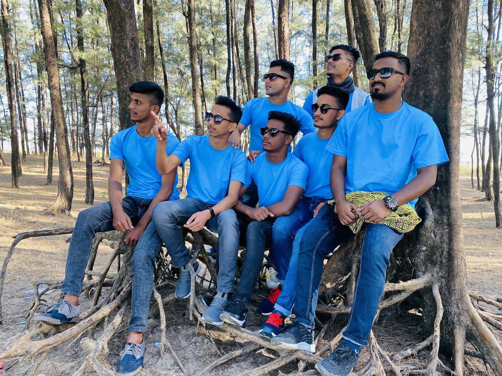Eight young men in matching bright-blue t-shirts sitting together on tree roots in a park