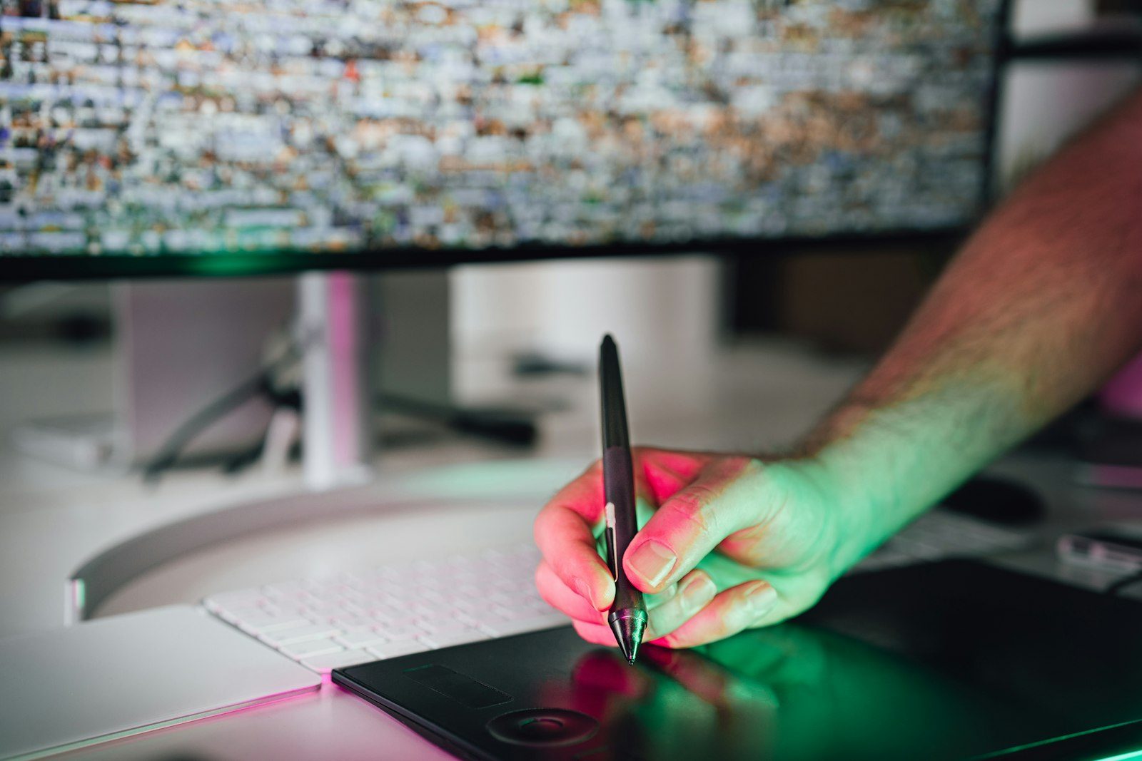 A hand holding a stylus over a drawing tablet in front of a large monitor, lit in green and pink
