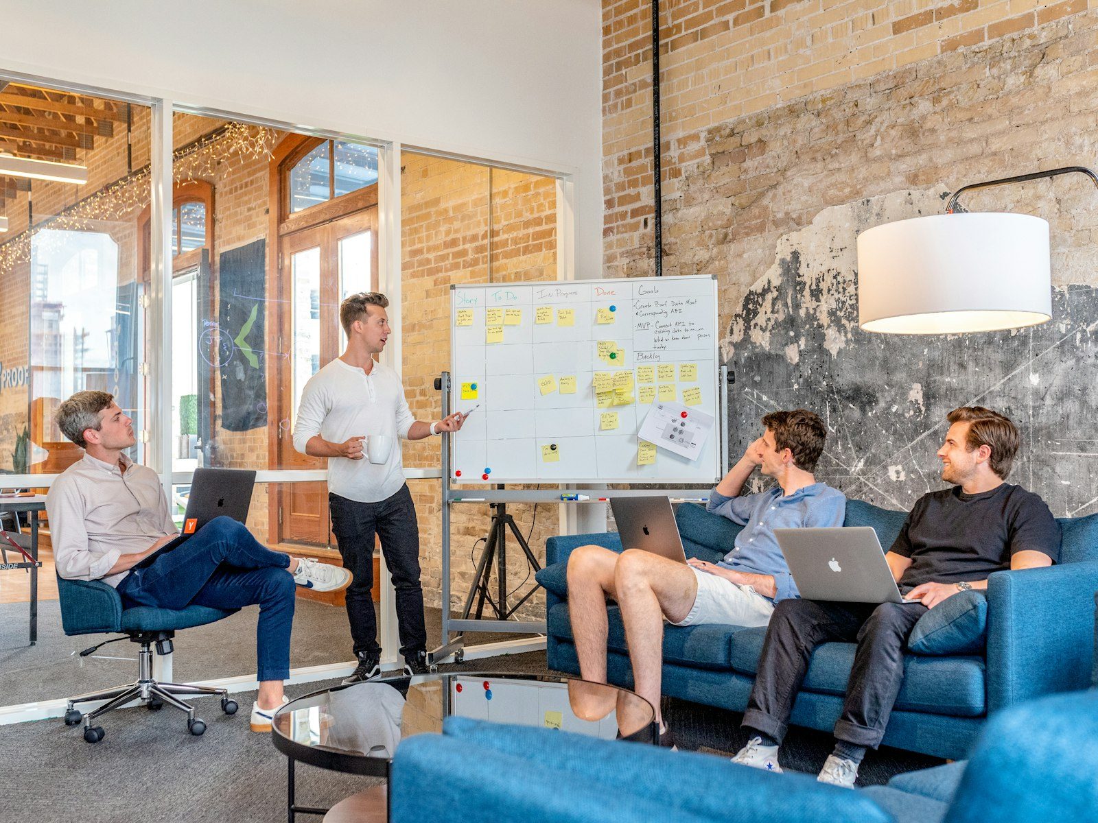 Four people in a brick-walled loft office, one standing at a whiteboard covered in sticky notes while the others listen