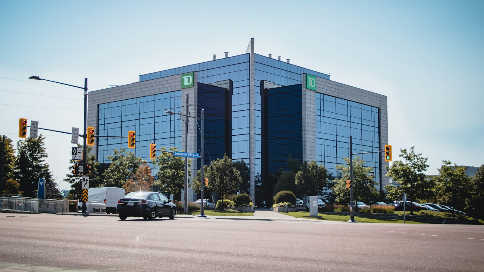 A blue-glass mid-rise office building on a suburban corner at a traffic-light intersection, with trees and a passing car