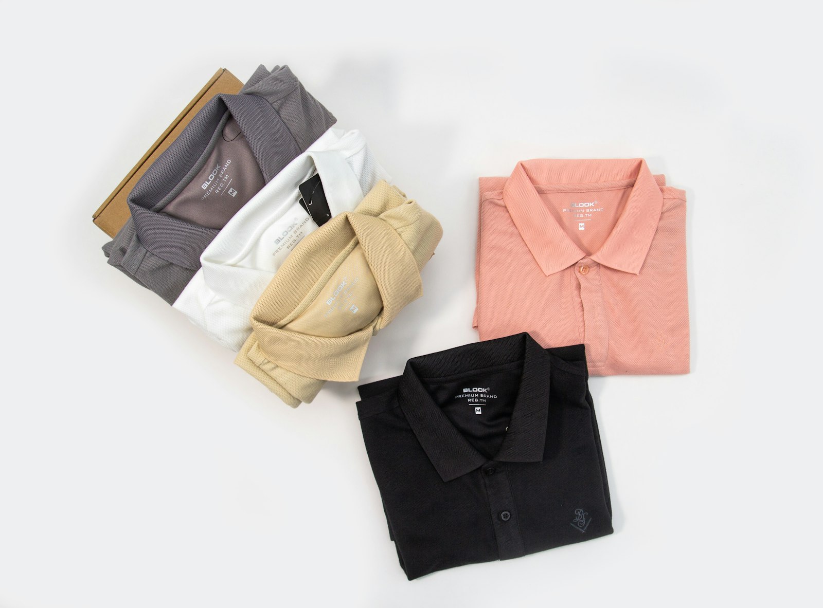 Five folded polo shirts in black, grey, white, tan and coral arranged flat on a white surface