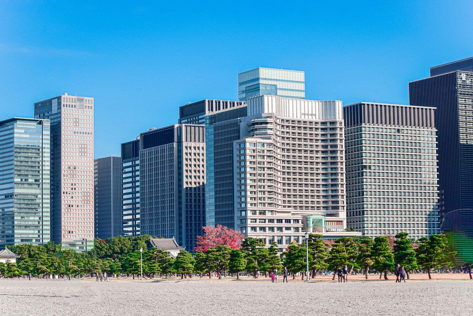 A cluster of glass and stone office towers of different heights behind a row of trees, with people crossing a wide plaza