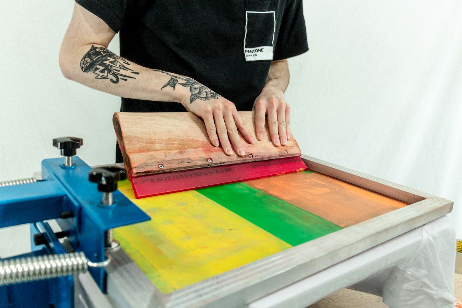 A printer in a black t-shirt pulling a squeegee across a screen on a manual press, with green and yellow ink on the mesh
