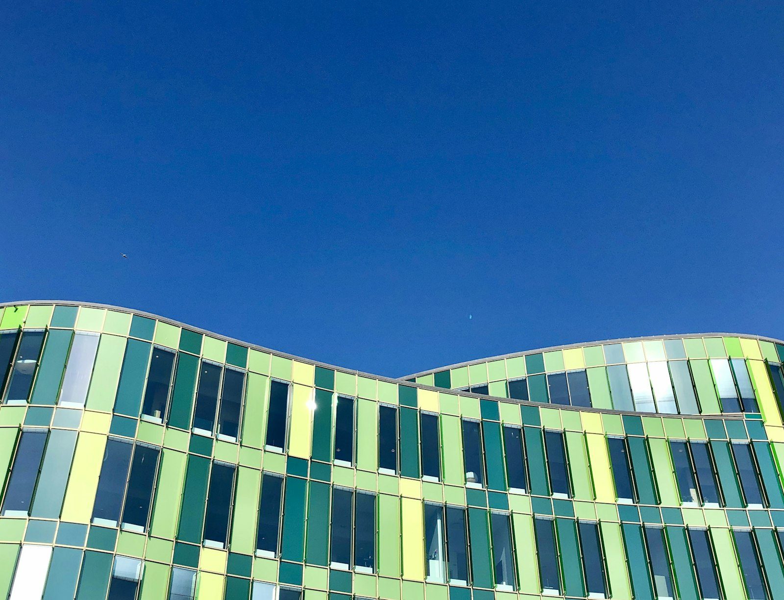 A modern mid-rise office building with a wavy facade of green, white and blue glass panels under a clear sky