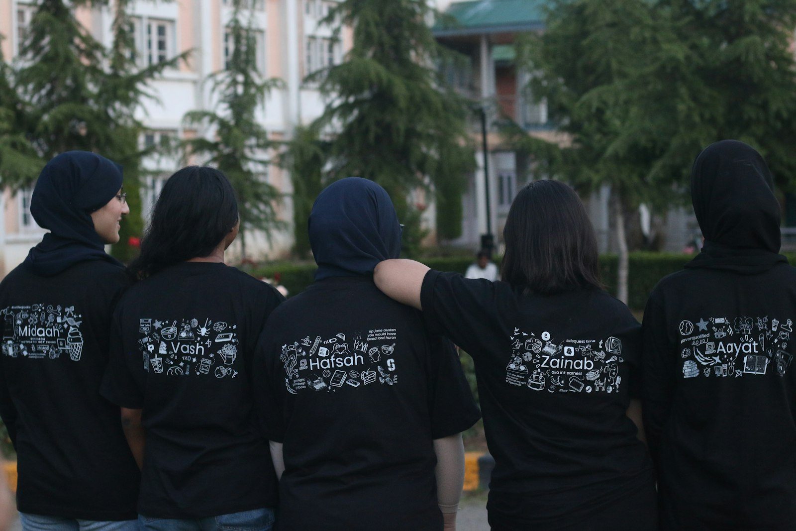 Five students seen from behind in matching black hoodies and t-shirts with a printed back design, standing in front of a school building
