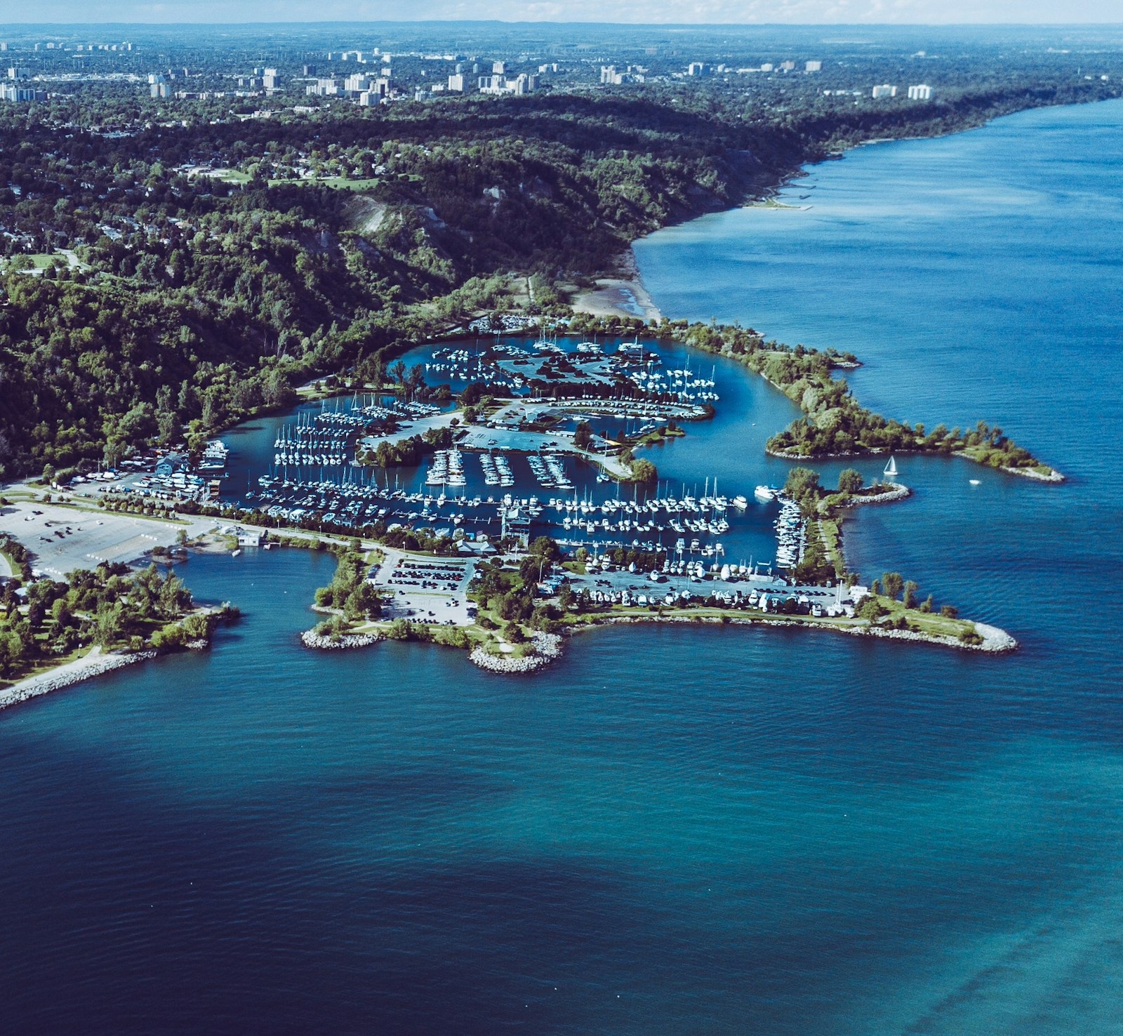 Aerial view of a marina full of boats at the foot of tree-covered bluffs on Lake Ontario on a bright day