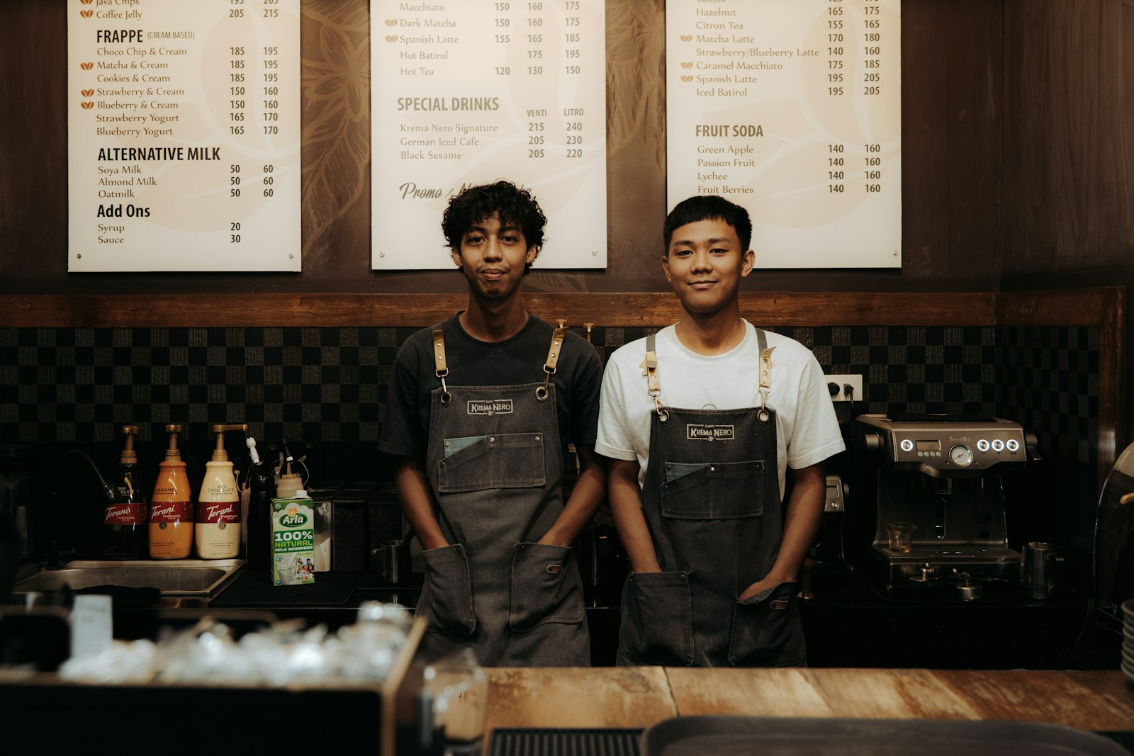 Two baristas in grey aprons standing behind a cafe counter under a chalkboard menu