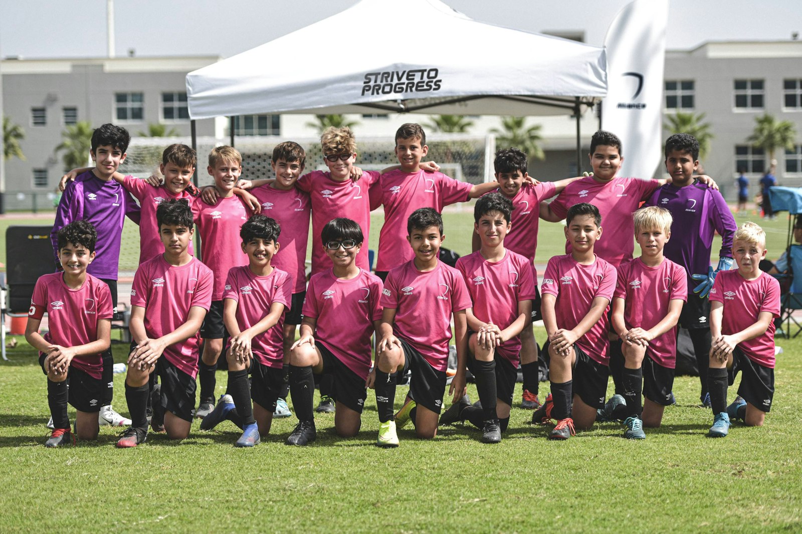A youth soccer team in matching pink jerseys and black shorts posing in rows on a field under a white tent