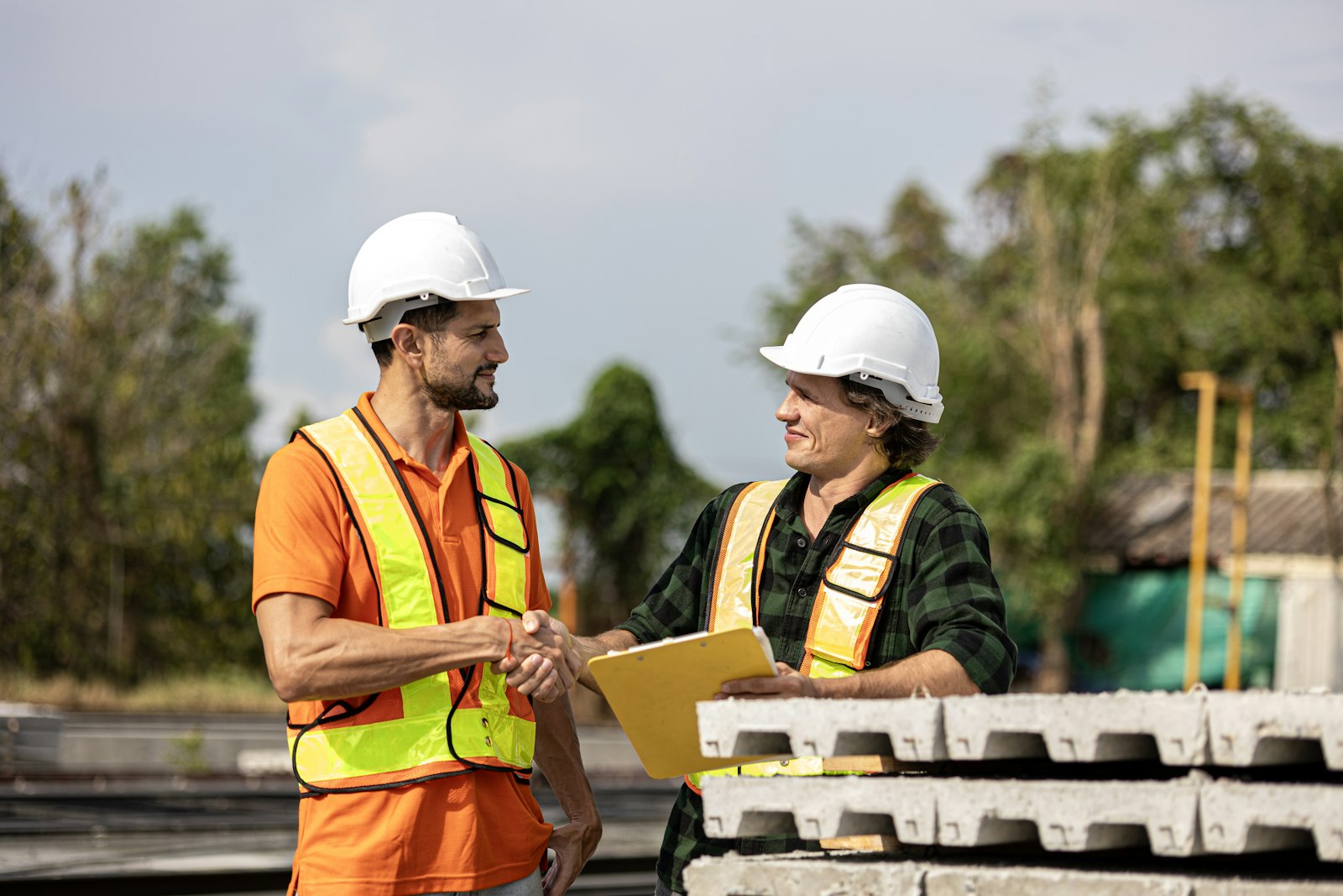 Two construction workers in white hard hats and orange and yellow hi-vis vests shaking hands over a clipboard beside stacked precast concrete